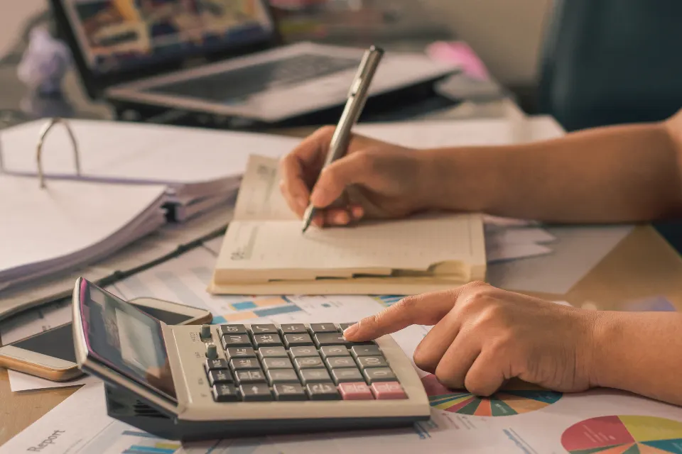 A hand pressing a desk calculator while another hand writes in a notebook, with papers and a laptop behind