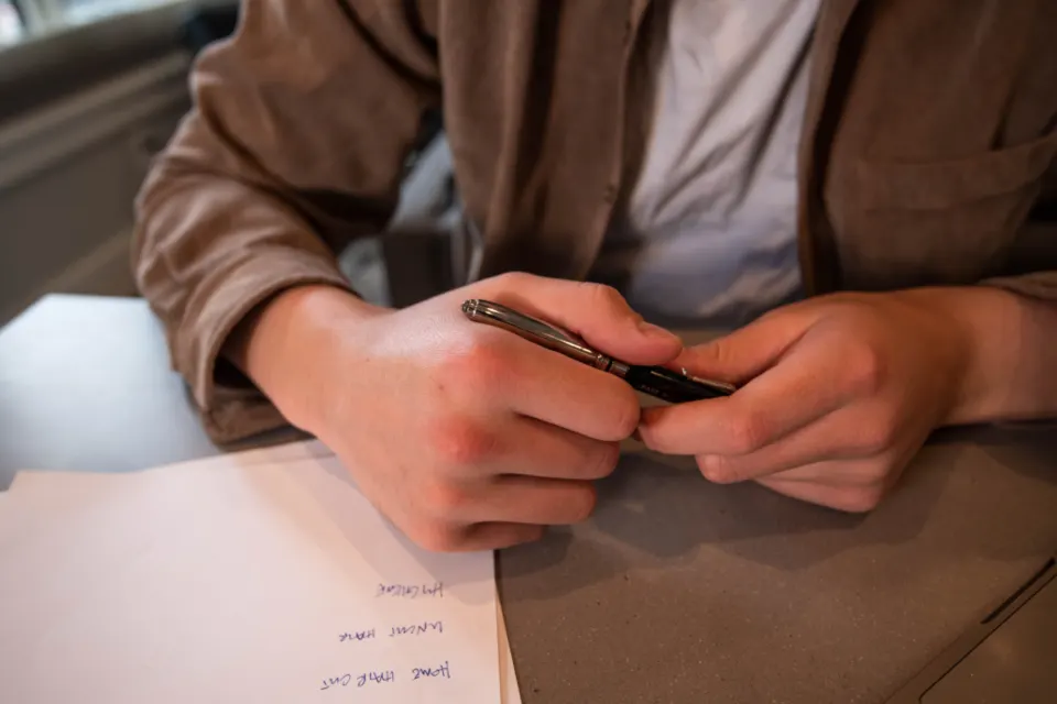 A person in a brown jacket at a desk holding a pen over a sheet of handwritten paperwork