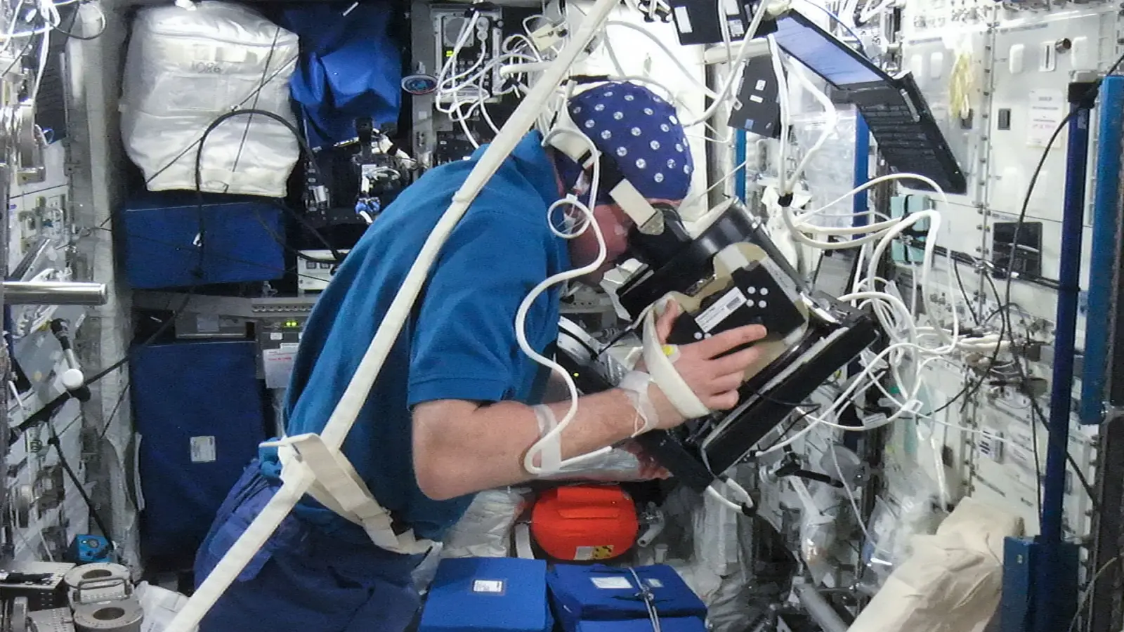 A person in a blue fabric cap dotted with white EEG electrodes, cables running from the cap, leans into a dark viewing device to watch a screen during a brain-wave recording