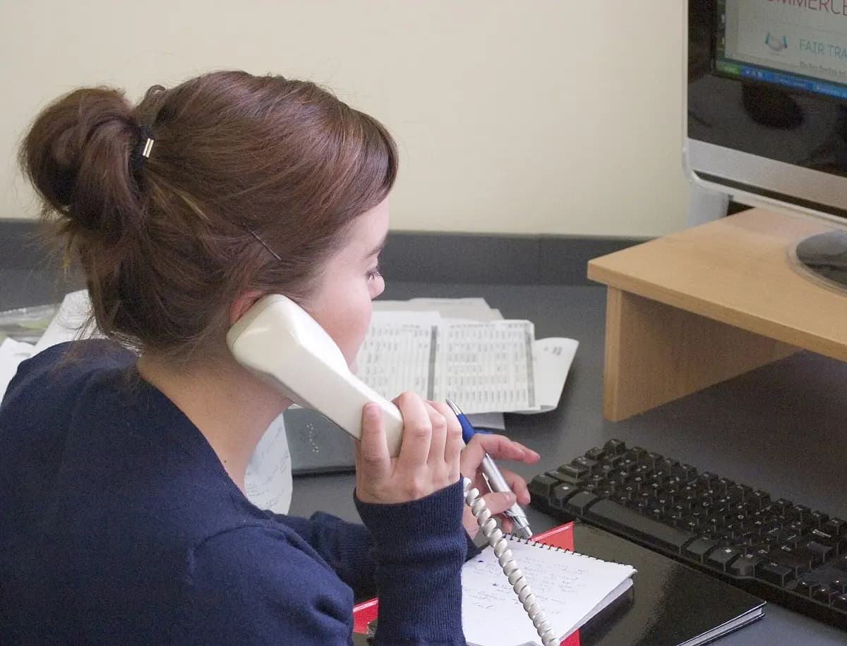 A woman at an office desk holding a telephone receiver to her ear, with a notebook and keyboard in front of her