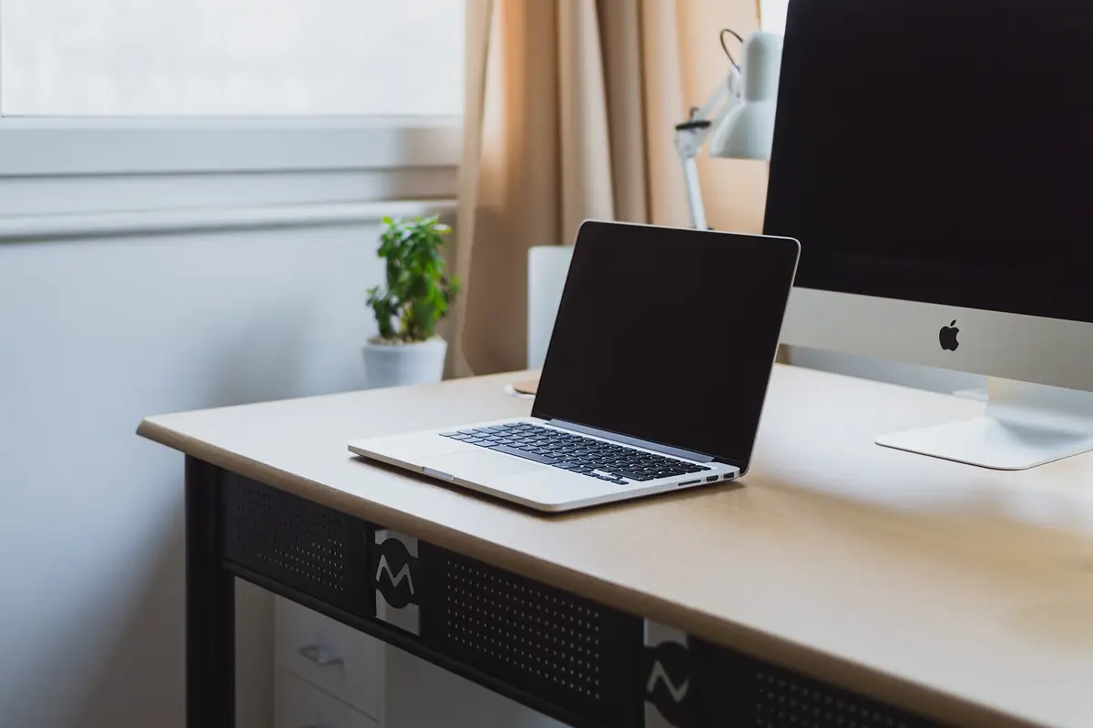 An open laptop on a tidy desk beside a window, with a small plant and a desktop monitor