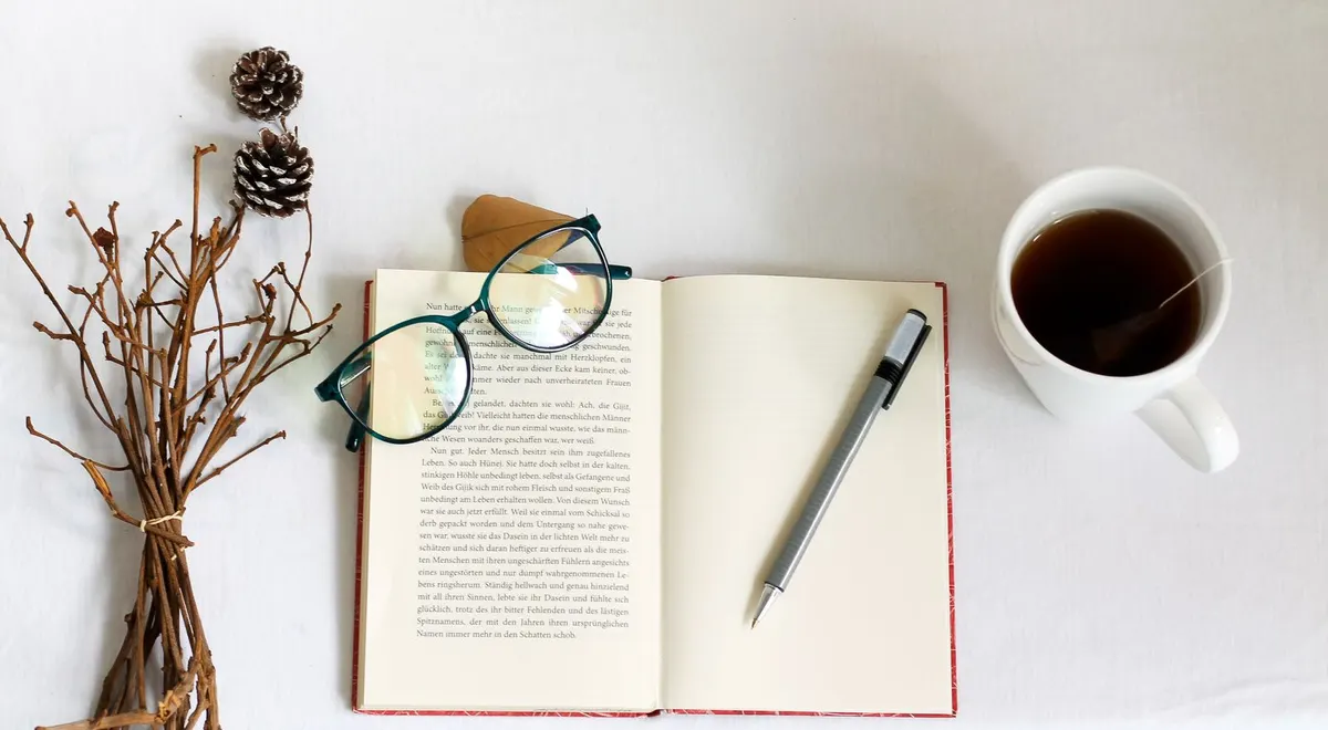 An open book with a pen, reading glasses and a cup of tea on a white table