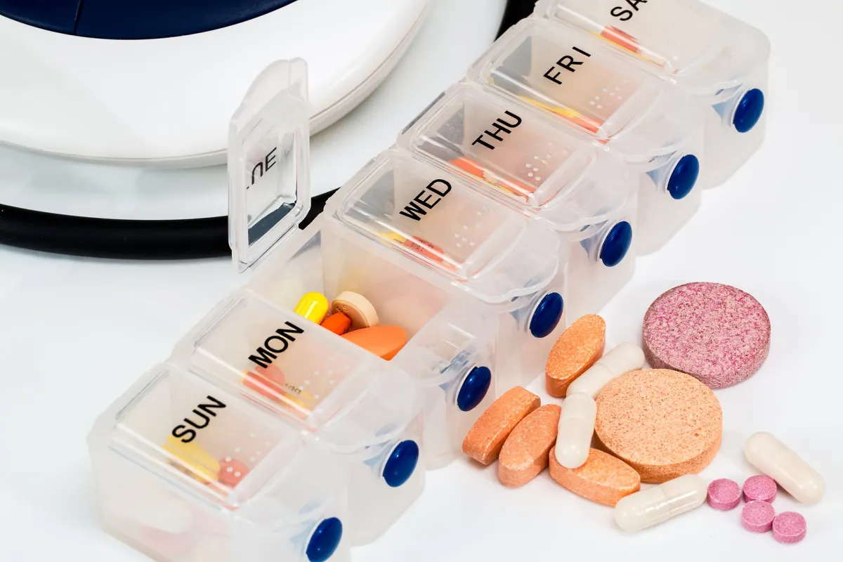 A clear weekly pill organizer with compartments marked SUN to SAT, some holding colored tablets and capsules, beside a scatter of orange, pink and white pills on a white table