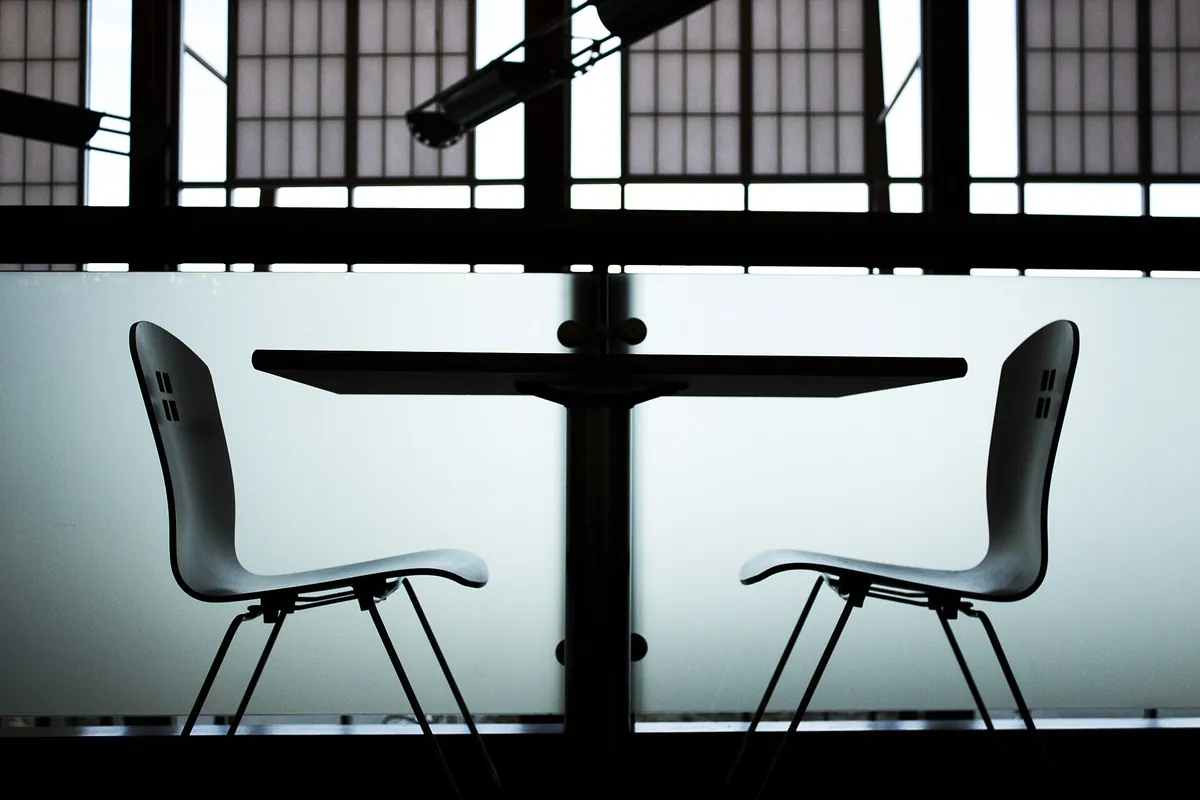 Two empty black chairs facing each other across a small round table, in silhouette against a bright window