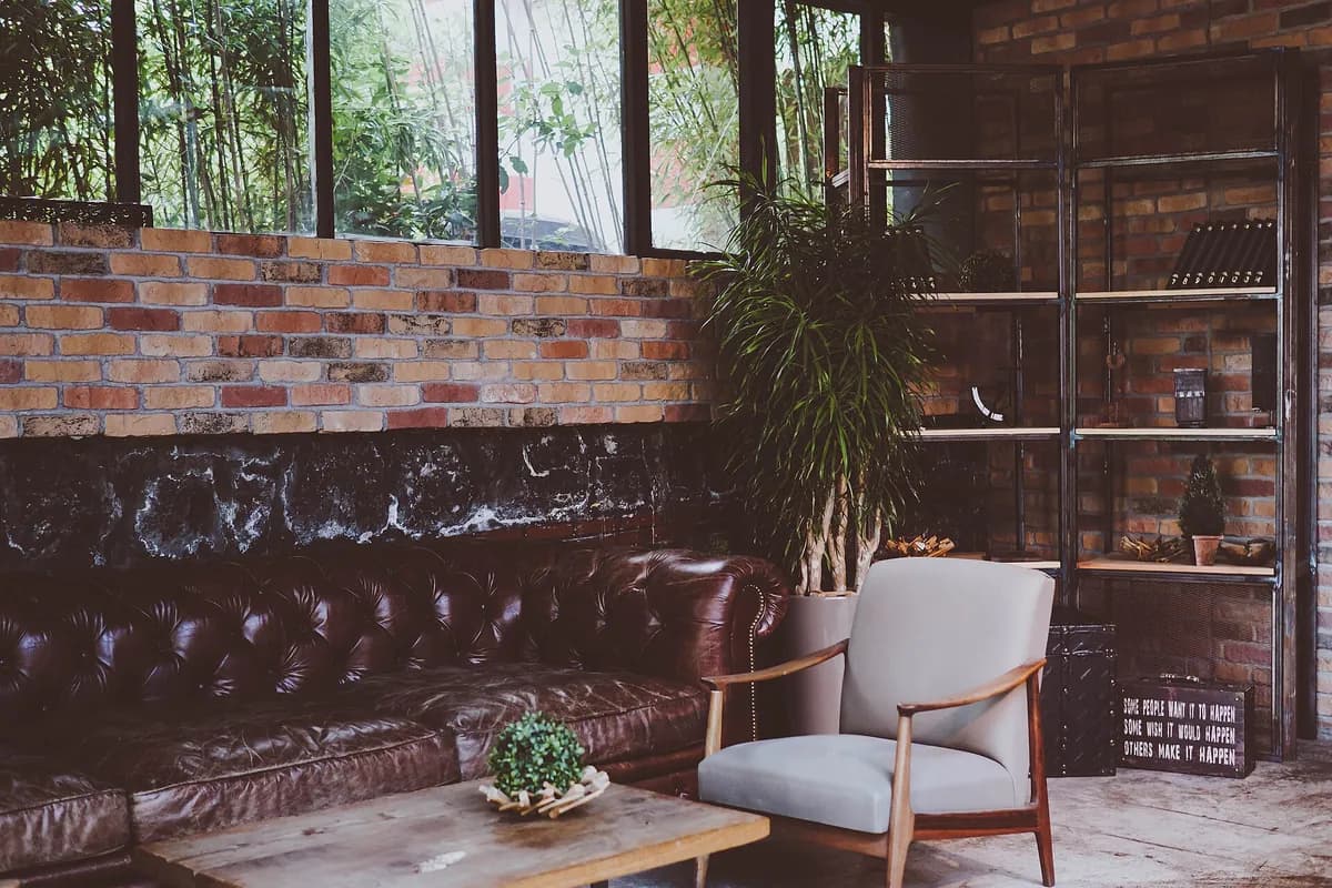 A quiet sitting room with a dark leather sofa, a light grey armchair, a low wooden table with a small plant, and a brick wall below tall windows
