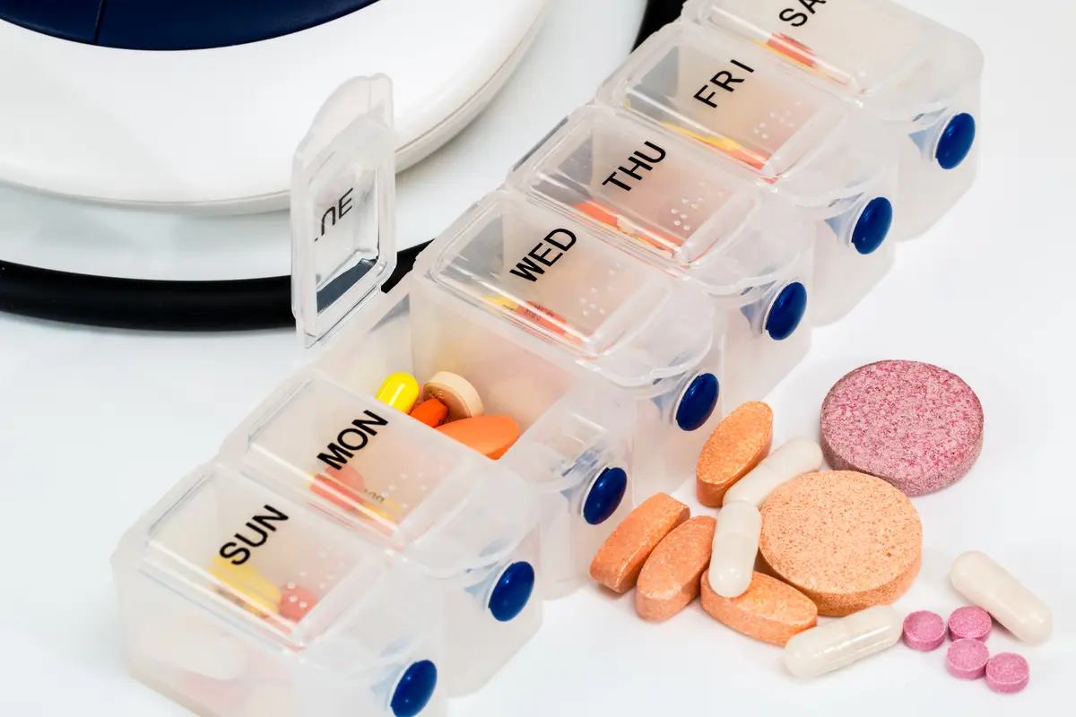 A clear plastic weekly pill organizer with boxes marked Sunday to Saturday, the Monday and Tuesday lids open, with assorted tablets and capsules spilled beside it